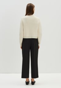 Crème trui met een geribbelde zoom, gecombineerd met hoge zwarte wijde broek en zwarte instappers. Effen achtergrond.