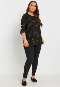 Svart texturerad tröja med gula och gröna mönster, vida ärmar och dekorativa knappar, ihop med svarta åtsittande byxor och loafers.