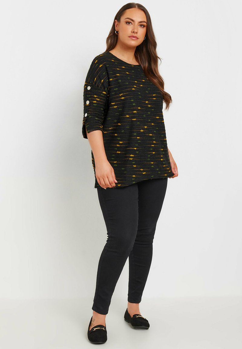 Svart texturerad tröja med gula och gröna mönster, vida ärmar och dekorativa knappar, ihop med svarta åtsittande byxor och loafers.