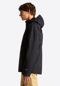 Ung mand iført sort hættetrøje med "DRYVENT" label, stående i profil, iført beige bukser mod en ensfarvet hvid baggrund.