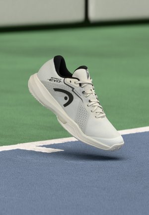 Weißer Sportschuh mit strukturiertem Obermaterial, schwarzen Akzenten und einer geformten Sohle, schwebend in der Luft über einer blauen Spielfeldoberfläche.