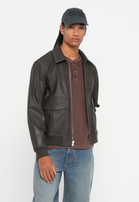 Nuori mies, jolla on pitkät letit, mustassa lippiksessä, tummanruskeassa vetoketjullisessa takissa, jossa on taskut, viininpunaisessa henley-paidassa ja sinisissä farkuissa, valkoista taustaa vasten.