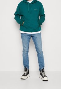 Sudadera verde azulado con bolsillo frontal y capucha, llevada sobre una camiseta blanca. Unos vaqueros azul claros y unas zapatillas negras de caña alta completan el atuendo.