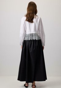 Cămașă albă cu dantelă la talie peste o fustă neagră, lungă până la podea. Are mâneci lungi și o textură fină. Design simplu.
