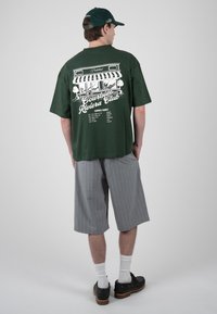 Homme portant une casquette vert foncé et un t-shirt vert foncé oversize avec un motif de café blanc et un short rayé gris à hauteur des genoux, debout de dos.
