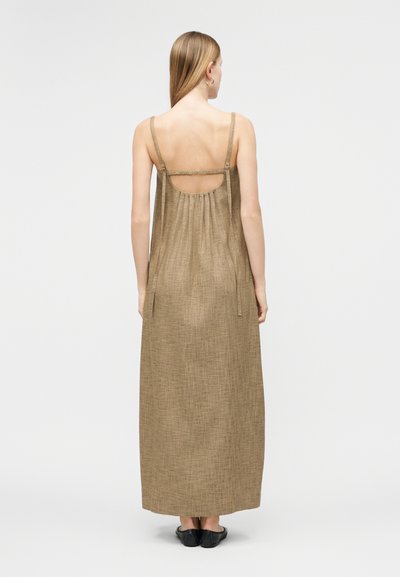 Γυναίκα με μακριά ξανθά μαλλιά, που φοράει μπεζ αμάνικο φόρεμα μέχρι τον αστράγαλο με λεπτές τιράντες και μαύρα επίπεδα παπούτσια, στεκόμενη πλάτη προς το μέρος μας.