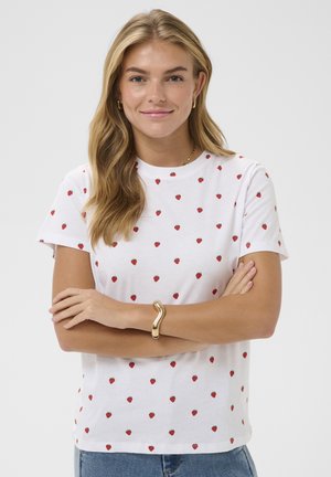 Tânără cu păr blond lung, purtând un tricou alb cu imprimeu de căpșuni, brățară aurie și cercei, stând cu brațele încrucișate.