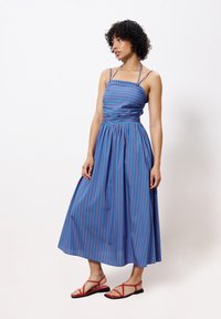 Blau- und rot gestreiftes, ärmelloses Maxikleid mit tailliertem Oberteil und schwingendem Rock, kombiniert mit roten flachen Sandalen.