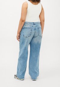 Personne portant un jean large bleu clair, un haut blanc sans manches et des chaussures de sport blanches, debout de dos sur un fond uni.