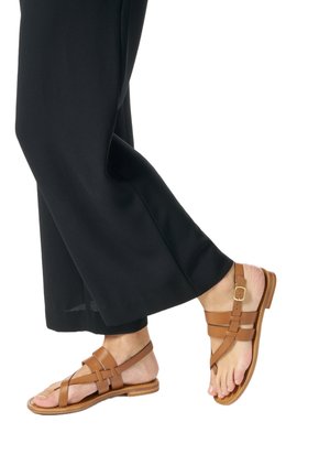 Persona che indossa pantaloni neri a gamba ampia e sandali piatti in pelle marrone con cinturini e fibbie, in piedi con un piede leggermente sollevato.