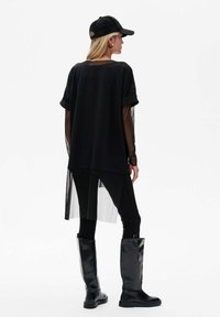 Vrouw in een zwarte doorschijnende lange mouwen top onder een losse zwarte T-shirt, zwarte legging, hoge zwarte laarzen en een zwarte baseball cap, van opzij gezien.