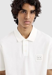Witte polo shirt van textiel met structuur, voorzien van een kraag, een knoopsluiting met drie knopen en een logo patch op de linkerkant van de borst.