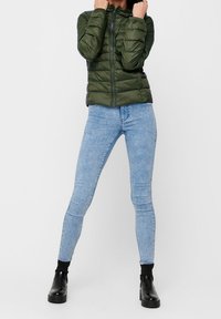 Doudoune vert olive avec une fermeture éclair à l'avant, des poignets côtelés et une capuche, associée à un jean skinny bleu clair et des bottines noires.