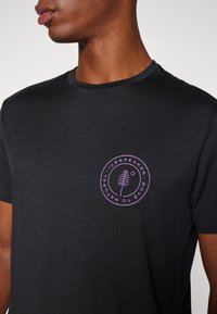 Svart kortermet t-skjorte i mykt materiale med et sirkulært lilla logo med trekdesign på brystet. Enkel rund hals-konstruksjon.
