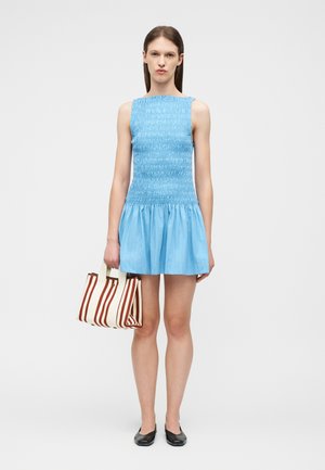 Mladá žena v modrých šatách bez rukávov s nariasením, držiaca bielu a hnedú pruhovanú tašku, stojaca pred jednoduchým svetlým pozadím.