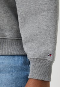 Harmaa huppari, jossa on resorit hihoissa ja helmassa, pehmeä tekstuuri, jossa on pieni Tommy Hilfiger -logopatchi punaisena, valkoisena ja sinisenä.