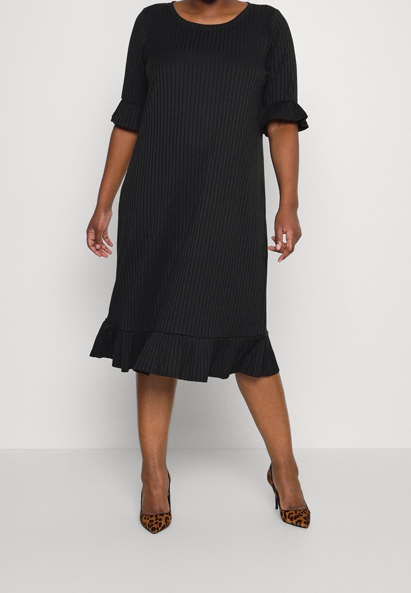 Femme portant une robe noire côtelée longueur genou avec manches et ourlet à volants, associée à des chaussures à talons hauts imprimé léopard.