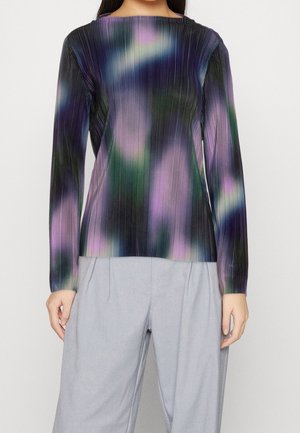 Kobieta ubrana w plisowaną bluzkę z długim rękawem z rozmytym wzorem w kolorach fioletu, zieleni i czerni, zestawioną z jasnoszarymi spodnie z szerokimi nogawkami.