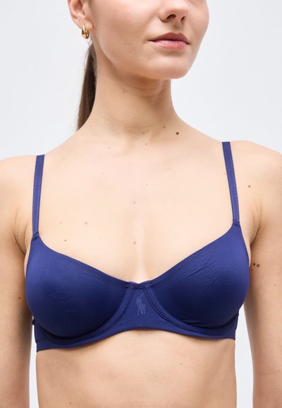 Γυναίκα που φοράει σουτιέν σε σκούρο μπλε χρώμα με μπανέλα, λεπτές τιράντες και μικρό κεντημένο λογότυπο στη μέση του μπροστινού μέρους, μπροστά σε απλό ανοιχτό φόντο.