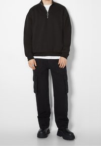 Svart zip-up sweatshirt med hög krage, vardera med löst sittande svarta cargobyxor med sidofickor. Matta svarta ankelskor fulländar looken.