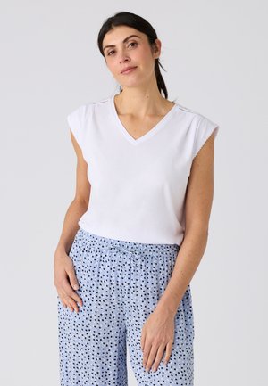 Mujer con el cabello oscuro recogido lleva una camiseta blanca sin mangas con cuello en V y pantalones sueltos azul claro con puntos negros y blancos.