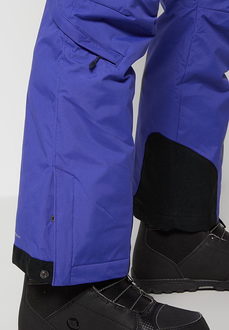 Pantaloni da neve viola con una tasca zip sul coscia, orlo nero rinforzato e tessuto impermeabile, abbinati a stivali da neve neri.