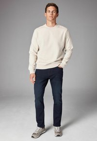 Sudadera de cuello redondo en color beige claro combinada con jeans oscuros y zapatillas grises. Diseño simple, textura lisa y puños elásticos.