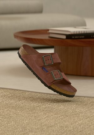 Καφέ σουέντ σανδάλι με δύο ρυθμιζόμενες αγκράφες και φελλό σόλα που αιωρείται πάνω από μια υφασμάτινη μπεζ μοκέτα σε ένα σαλόνι.