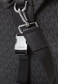 Tissu noir à motifs avec quincaillerie en métal argenté. Dispose d'une boucle à pression et d'une sangle texturée, offrant un design fonctionnel et durable.