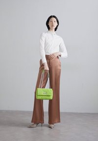 Pantaloni a gamba larga in satin marrone, camicia bianca con colletto, in mano una borsa trapuntata verde brillante con hardware argentato. Indossati con tacchi alti beige.