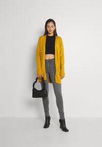 Cardigan largo mostaza con puños acanalados, top corto negro, jeans skinny grises, bolso negro con asa retorcida y botines negros de tacón.