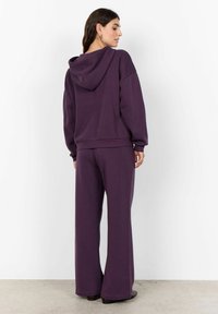 Sudadera morada de corte relajado y capucha con cordón, combinada con pantalones de pierna ancha a juego. Textura suave, diseño minimalista, sin patrones visibles.
