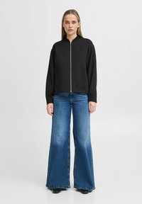 Svart zip-up jacka med hög krage, tillsammans med blå utsvängda jeans. Jackan har långa ärmar och en mjuk textur.