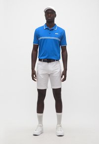 Άνδρας που φοράει μπλε πόλο μπλούζα, λευκά σορτς, λευκή ζώνη, λευκές κάλτσες, λευκά αθλητικά παπούτσια και λευκό καπέλο, στέκεται μπροστά σε απλό φόντο.