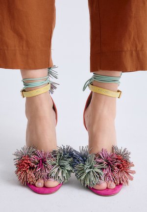 Pies con sandalias rosas de punta abierta con adornos de pompones multicolores y tiras amarillas y verde menta en los tobillos, combinados con pantalones color óxido.