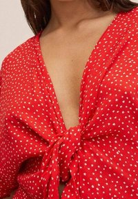 Femme portant un haut rouge à petits pois blancs, avec un profond décolleté en V et un nœud noué à l'avant sur la poitrine.
