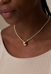 Auksinis ir kristalų choker tipo kaklo papuošalas su centriniu auksiniu ašaros formos pakabuku, aplink kurį yra mažų apvalių kristalų grandinėlė. Matyti tekstūruota oda.