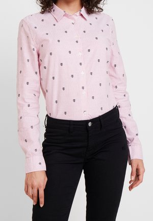 Kobieta w różowo-białej koszuli w paski zapinanej na guziki, ozdobionej małymi czarnymi nadrukami pszczół, w połączeniu z czarnymi dopasowanymi spodniami.