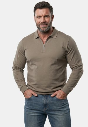Mand med kort mørkt hår og skæg iført en taupe langærmet trøje med lynlås og blå jeans, stående med hænderne i lommerne og smilende.