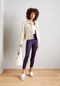 Beige kort jasje met twee voorzakken, witte tanktop, marineblauwe leggings en witte hoge muilen, gecombineerd met een witte handtas.