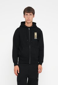 Black zip-up hoodie made of cotton, featuring a front pocket, drawstrings, and a gold graphic on the left chest area. Rubber logo accent.