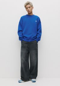 Sweatshirt oversize bleu avec des poignets côtelés, associé à un jean noir à jambes larges et des baskets bleu et blanc. Détail logo simple sur le t-shirt.