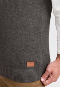 Grauer Strickpullover mit geripptem Saum und einem Lederetikett mit der Aufschrift "BLEND" nahe dem unteren Rand. Der Stoff wirkt weich und strukturiert.