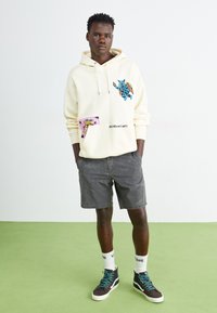 Creme hoodie med livlige blå og pink grafiske prints, parret med grå shorts og sorte sneakers med teal detaljer.
