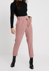 Rose-colored high-waisted trousers with pleats and a buckle belt, made from a smooth fabric, paired with a fitted black top and shiny black boots.