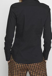 Zwarte nauwsluitende shirt met een kraag en knoopmanchetten, gemaakt van gladde stof. Draag het met een geruite broek in oranje en zwart.