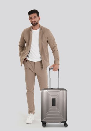 Maleta rígida de color gris metálico, con un diseño estriado y un asa negra, colocada al lado de una persona vestida de manera casual con atuendo beige.