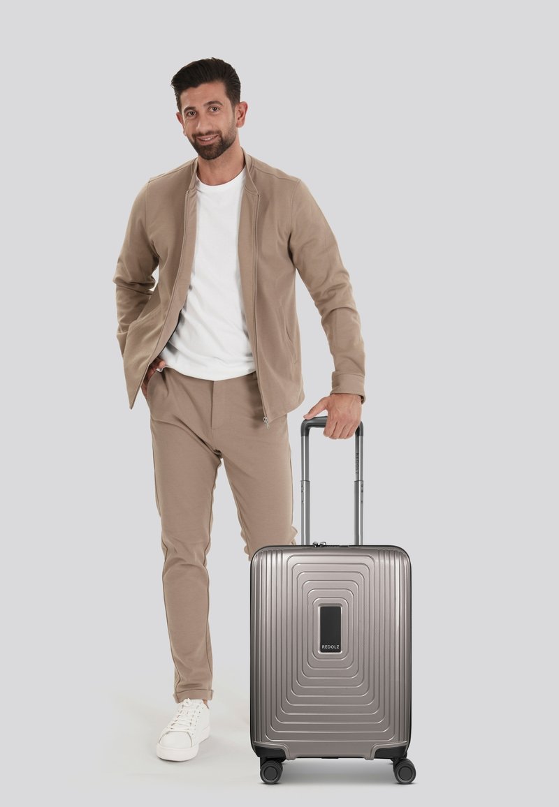 Maleta rígida de color gris metálico, con un diseño estriado y un asa negra, colocada al lado de una persona vestida de manera casual con atuendo beige.
