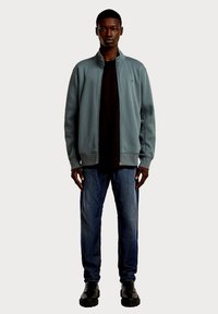 Jasnoniebieska kurtka zapinana na zamek, noszona na czarnym t-shircie, w połączeniu z ciemnoniebieskimi dżinsami i czarnymi butami. Kurtka ma luźny krój i wysoki kołnierz.