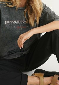 Černé vybledlé tričko s potiskem "Revolutionary American Vintage" a designem americké vlajky; spárováno s volnými černými kalhotami a sandály.